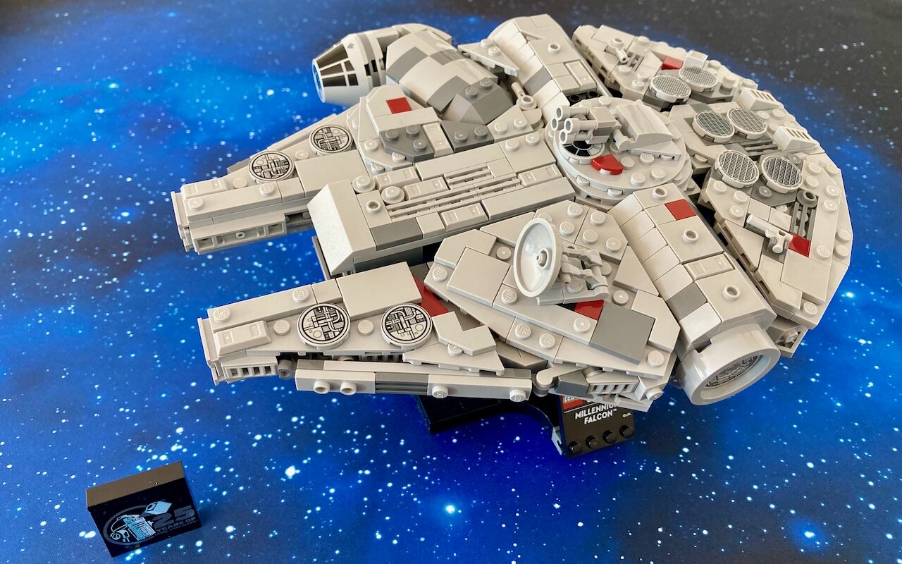 Das Foto zeigt ein LEGO Modell des Millennium Falcon, das auf einer Oberfläche mit Sternenmuster steht, was den Eindruck eines Fluges durch den Weltraum vermittelt. Im Vordergrund ist. Das Modell selbst ist detailliert, mit grauen und weißen Teilen sowie roten Akzenten.